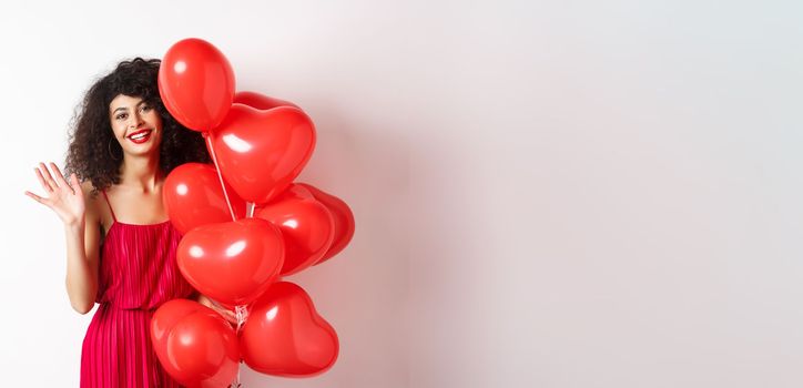 Beautiful lady with curly hair, standing near valentines holiday balloons and waving hand, saying hello, standing against white background.