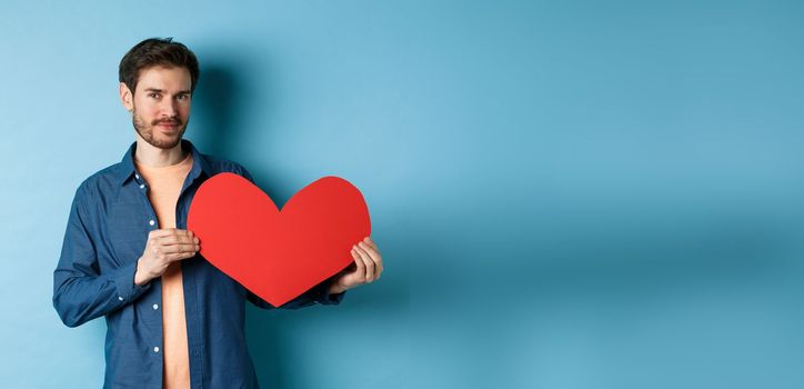 Handsome bearded guy showing valentines day cutout and smiling, standing in casual clothes over blue background. Copy space