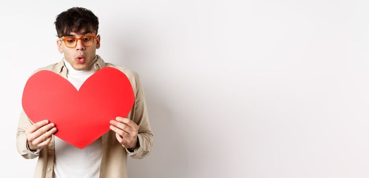 Surprised handsome man receive big red heart postcard on Valentines day, looking at gift with amazement, enjoying lovers day, standing over white background.