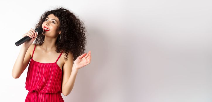 Beautiful lady in red dress singing songs in microphone, smiling and looking up, standing on white background.