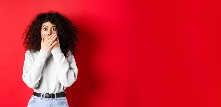 Image of speechless caucasian woman covering mouth with hands, looking shocked at camera, standing in casual clothes on red background.