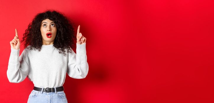 Impressed european female model in casual clothes, checking out promo with dropped jaw, looking and pointing fingers up, showing logo, standing on red background.