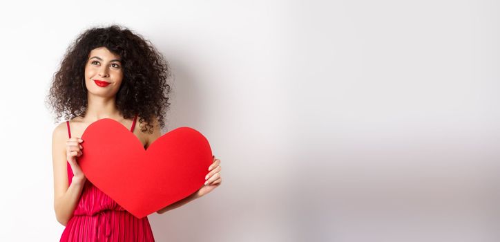 Young romantic woman dreaming about love on Valentines day, looking for soulmate, holding big red heart cutout and gazing pensive left, white background.