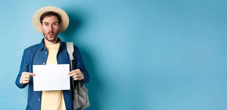 Excited tourist in straw hat, holding empty piece of paper and looking amused, going on summer travel, standing on blue background.