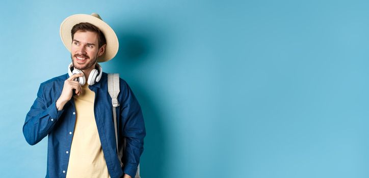 Handsome tourist with backpack and straw hat looking aside, smiling and touching headphones, standing on blue background.