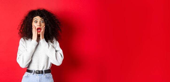 Portrait of shocked woman scream amazed, touching face and looking at camera at impressive promo offer, standing in sweatshirt on red background.