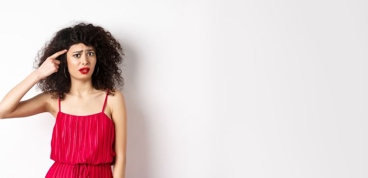 Shocked and confused caucasian woman in red dress scolding someone crazy, pointing finger at head and frowning, standing over white background.