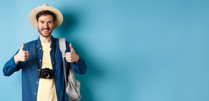 Cheerful handsome man recommending summer vacation place, showing thumbs up and smiling in approval. Tourist leave positive feedback, standing with camera and backpack on blue background.