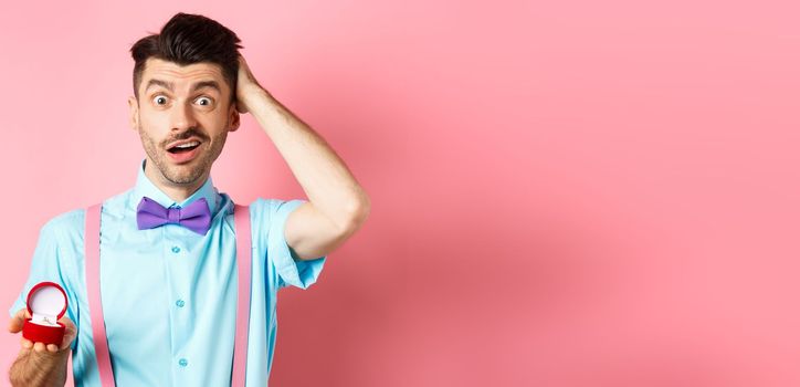 Valentines day. Confused funny guy standing with engagement ring, scratching head with unsure face, dont know what say, making proposal, standing over pink background.