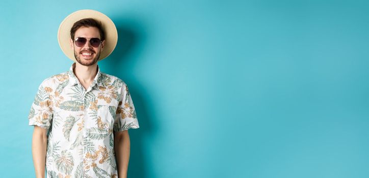 Handsome smiling guy in sunglasses and summer shirt, enjoying vacation on tour, standing on blue background.