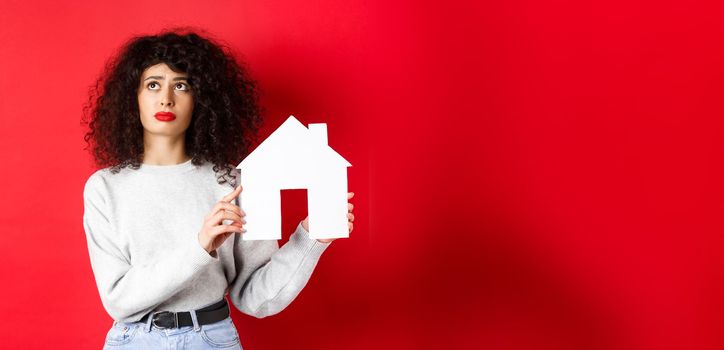 Real estate. Sad woman thinking of buying own house, showing paper home cutout and looking up with uspet face, standing on red background.