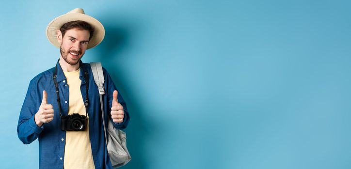 Handsome caucasian tourist in straw hat, holding backpack and photo camera, showing thumbs up, recommending travel agency or hotel resort, blue background.