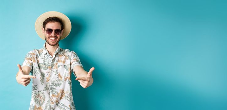 Happy tourist in summer hat and hawaiian shirt, pointing fingers at logo on center, showing something, standing on blue background.