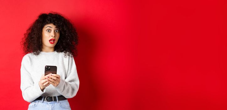 Image of pretty curly woman with shocked face, reading message on smartphone startled, standing against red background.