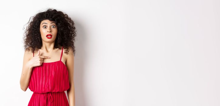 Surprised young woman in red dress pointing at herself, being confused and shocked, standing on white background. Copy space