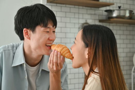 Cute young couple eating croissant while having breakfast in kitchen, enjoying leisure weekend time together.