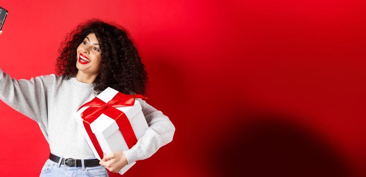 Holidays and tech concept. Happy woman taking selfie with her gift, holding present and smartphone, standing on red background.