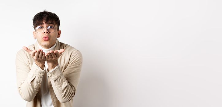 Cute boyfriend sending air kiss at camera on happy Valentines day, looking tender at lover, standing in casual clothes and glasses on white background.