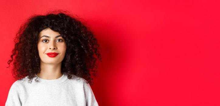 Close up portrait of beautiful modern woman with curly hairstyle and red lips, smiling happy at camera, standing on studio background.