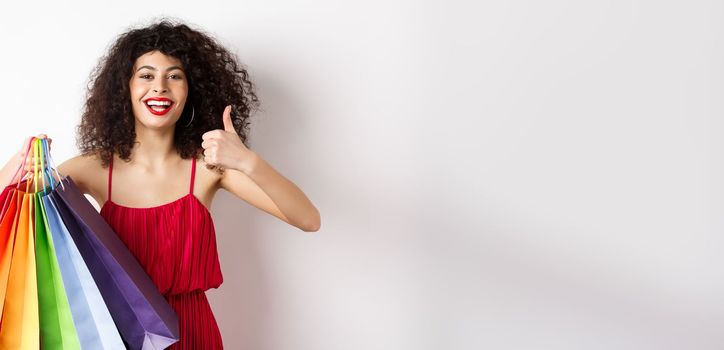 Fashionable woman in red dress going shopping, holding bags and showing thumbs up, recommend store, standing on white background.