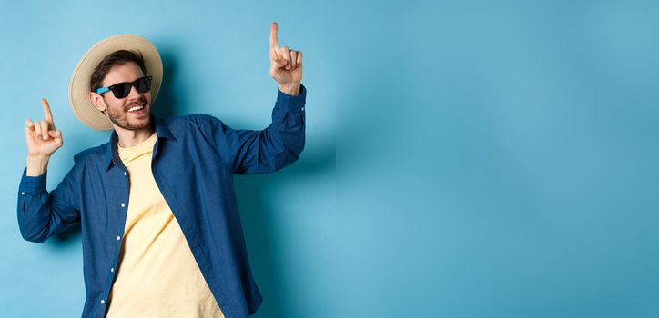 Funny tourist dancing on vacation, pointing fingers up, wearing summer hat and sunglasses, blue background.