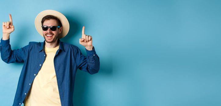 Cheerful caucasian guy in straw hat and sunglasses, dancing and having fun on vacation, standing over blue background. Concept of summer tourism.