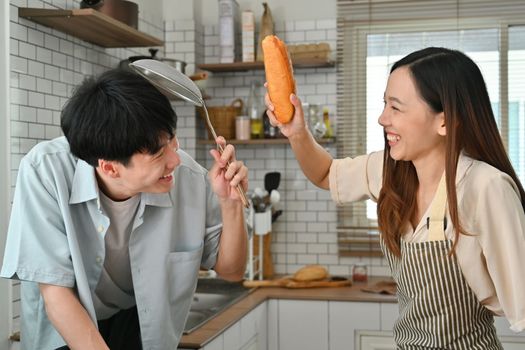 Overjoyed married couple using kitchenware and bread baguettes fighting in modern kitchen.