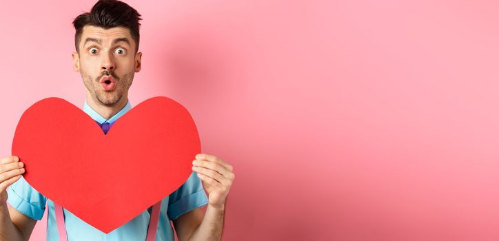 Valentines day concept. Amused young man with bow-tie and moustache, showing heart cutout and looking for love, standing on pink romantic background.