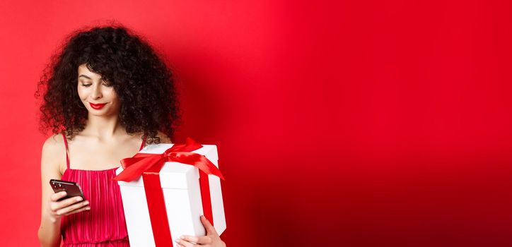 Valentines day and shopping. Beautiful caucasian woman in red dress, holding gift box from lover and using mobile phone, reading message on smartphone, studio background.