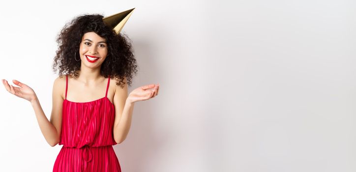 Beautiful caucasian woman in red dress, celebrating holiday, wearing party hat and smiling, standing on white background. Copy space