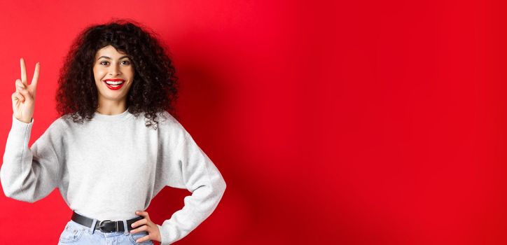 Young pretty lady in sweatshirt showing number two, making an order and smiling, standing on red background.