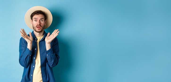 Image of shocked tourist in straw hat panicking, raising hands up and stare startled at camera, standing on blue background.