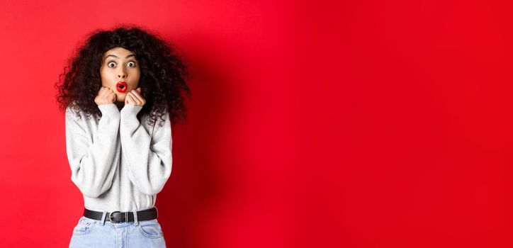 Surprised young woman looking with disbelief and amazement at camera, saying wow and standing in awe on red background.