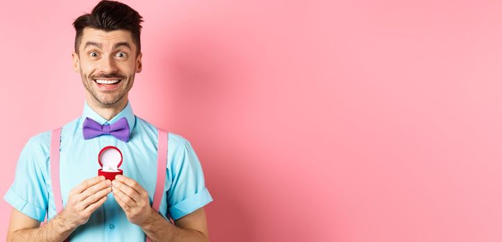 Valentines day. Romantic man in bow-tie showing engagement ring and smiling, asking to marry him, making proposal to lover on pink background.