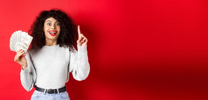 Young excited woman having an idea how make money, showing dollar bills and raising finger in eureka sign, standing on red background.