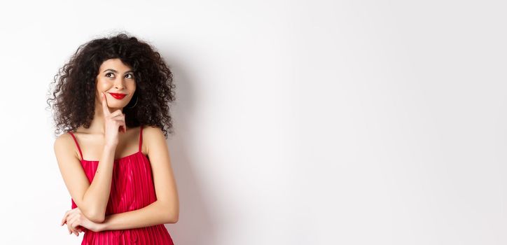 Image of elegant woman in red dress, looking pensive and smiling, thinking of romantic date, standing over white background.