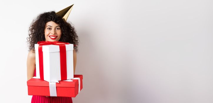 Holidays and celebration. Beautiful lady with curly hair, wearing party hat and holding big presents, smiling happy, receiving birthday gifts, standing on white background.