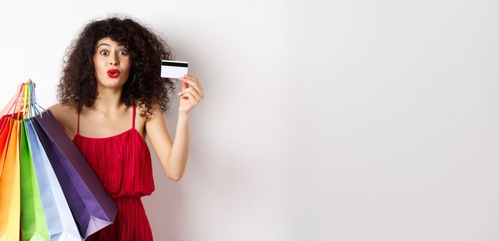 Excited woman in red dress, holding shopping bags and showing plastic credit card, shop with discounts, white background.