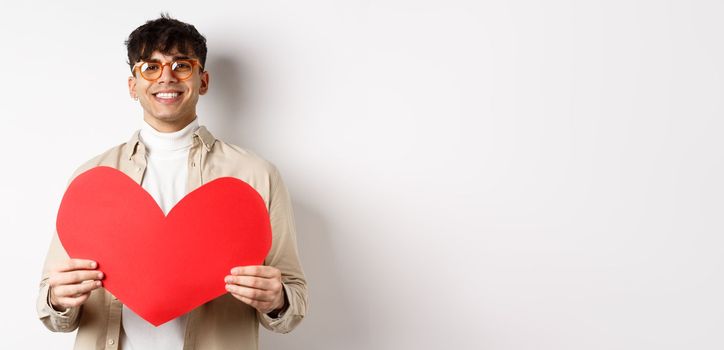 Attractive modern man smiling and looking at camera hopeful, holding big red Valentines heart, waiting for soulmate on lover day date, standing over white background.