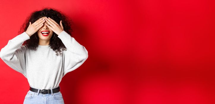 Happy beautiful woman with curly hair and red lips, covering eyes with hands and waiting surprise, smiling excited, standing against red background.