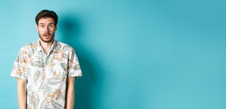 Image of shocked tourist guy drop jaw, gasping and looking startled, standing in hawaiian shirt on blue background. Copy space