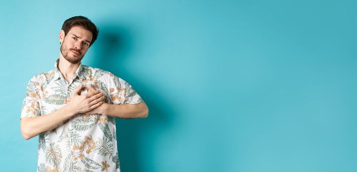 Passionate and romantic guy in hawaiian shirt, holding hands on heart and looking with sympathy, being in love, standing on blue background.