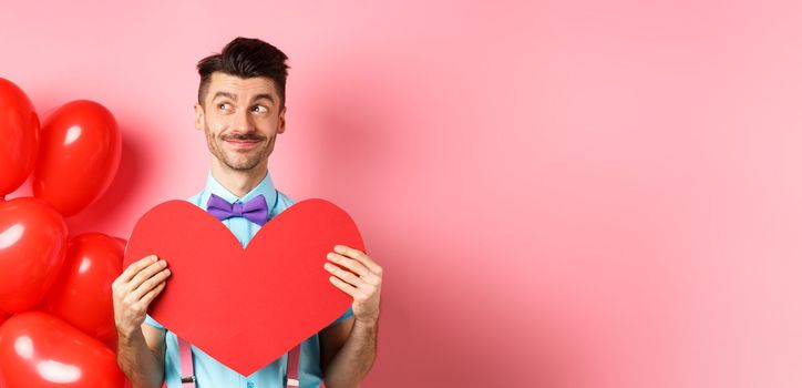 Valentines day concept. Smiling handsome man looking dreamy left, waiting for true love girlfriend with big red heart cutout, standing over pink background.