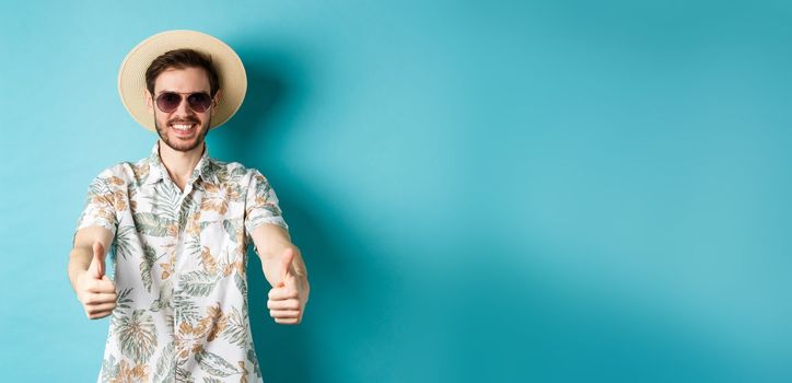 Happy tourist in sunglasses and straw hat showing thumbs up and smiling, enjoying summer holiday, standing on white background.
