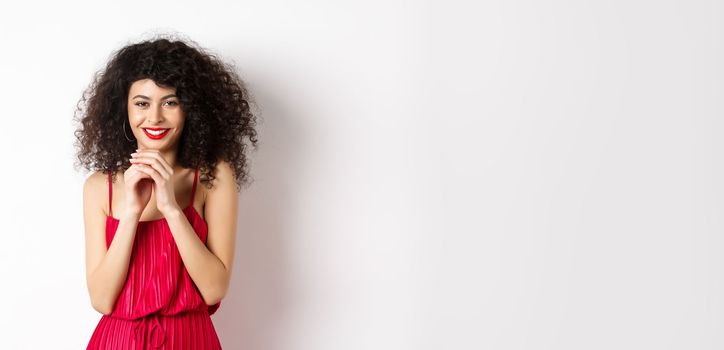 Cunning young woman with curly hair, steeple fingers and smiling devious, having a plan, standing in red dress on white background.