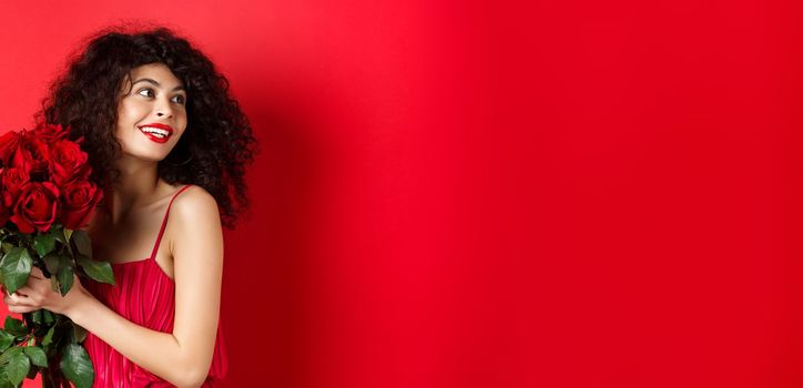 Tender young woman in elegant red dress, holding romantic bouquet of red roses and looking right, smiling dreamy, thinking about lover on Valentines day, studio background.