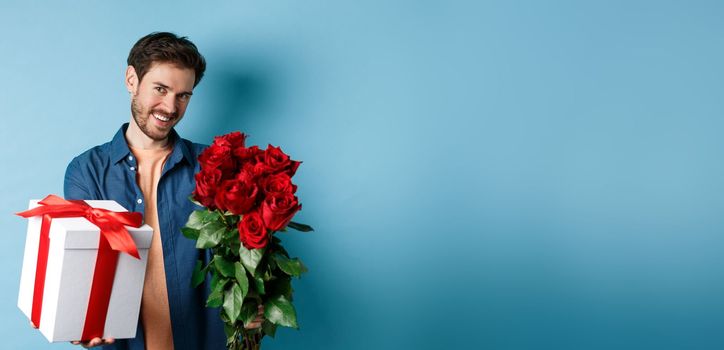 Love and Valentines day concept. Romantic smiling man giving you gift box and bouquet of flowers on date, standing over blue background.