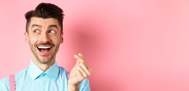 Valentines day concept. Romantic guy showing finger heart and looking left, smiling happy, standing on pink background dreamy.