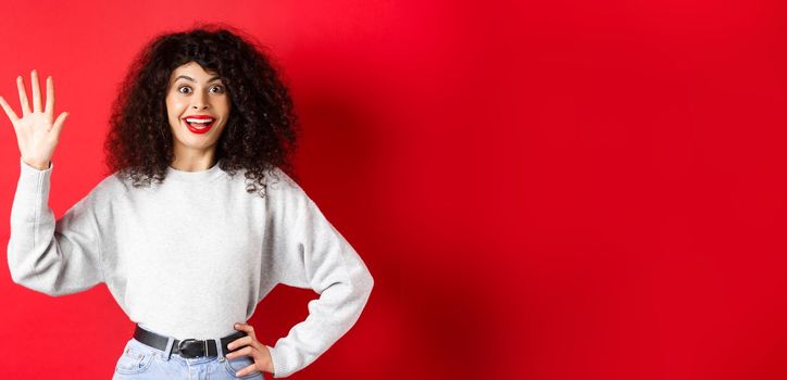 Excited woman with curly hair showing number five with fingers, making order, standing against red background.