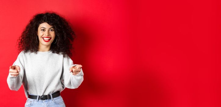 Cheerful woman with curly hair inviting you, recruiting newbies, pointing fingers at camera and smiling, praising good work, standing on red background.